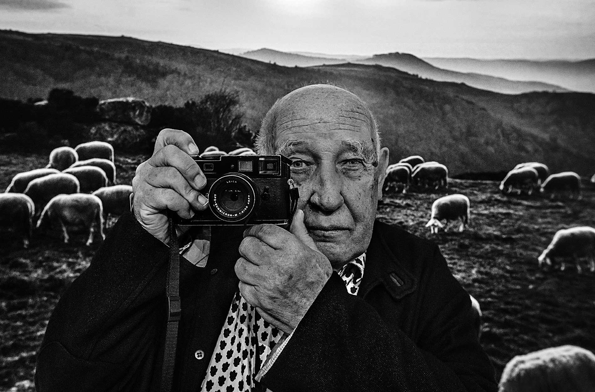 Raymond Depardon debout, appareil photo à la main, capturé par Eric Canto en coulisses, lumière naturelle